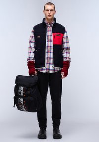 Chaleco de forro polar, en navy y rojo, sobre una camisa de cuadros. El modelo sostiene una mochila negra con logotipo. Unos pantalones negros y zapatos resistentes completan el atuendo.