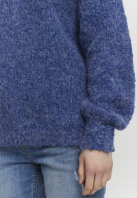 Pull en tricot bleu chunky avec une finition texturée et des manches légèrement ballons, associé à un jean délavé. Matériau doux et fuzzy.