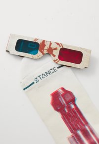 Gafas 3D con lentes rojos y azules, con un diseño de cartón texturizado, colocadas sobre calcetines blancos que muestran un patrón gráfico en rojo.