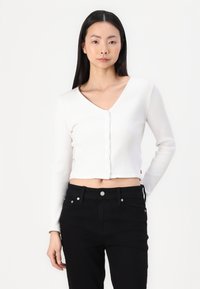 Λευκό, ριγωτό, κοντό ζακέτα με V-λαιμόκοψη, διαθέτοντας μια σειρά κουμπιών μπροστά και μακριά μανίκια. Συνδυασμένο με μαύρα τζιν.