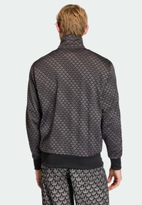 Čierna vzorovaná bunda s vysokým golierom, ktorá má textúrovaný dizajn prekrytých tvarov. Na lemovaní rukávov a spodnom okraji sú rebrované manžety, doplnená o nohavice rovnakého vzoru.