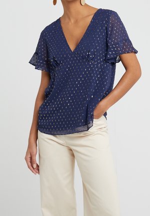 Mujer que lleva una blusa azul marino de manga corta con puntos plateados y pantalones anchos color crema, con la mano en el bolsillo, sobre fondo liso y claro.