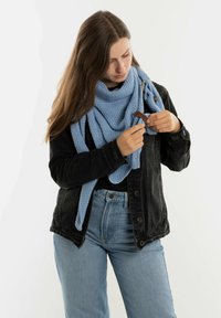 Lichtblauwe gebreide sjaal gedragen over een zwarte spijkerjack; model past een bruin accessoire aan in de handen. Zachte textuur, driehoekige vorm.