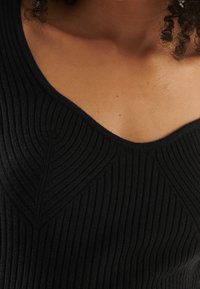 Haut noir en tricot côtelé avec un décolleté en V, présentant des motifs texturés en forme de feuilles sur la poitrine et un tissu lisse et doux.