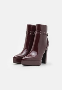 Bottines en cuir verni bordeaux avec un haut talon bloc, des orteils pointus et des brides à clous au niveau de la cheville. Fini brillant avec des accents texturés.