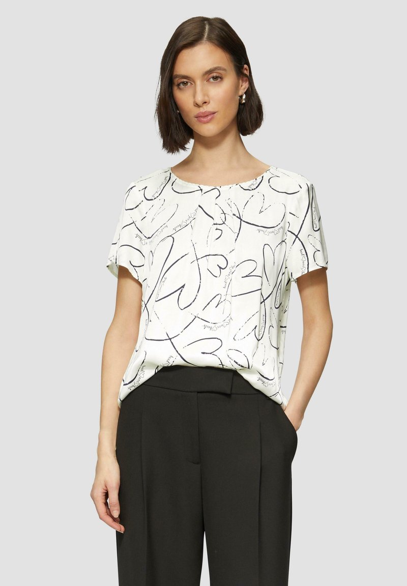 Blouse à manches courtes en tissu léger blanc avec des motifs de cœurs abstraits noirs et un col arrondi, associée à un pantalon noir taille haute.