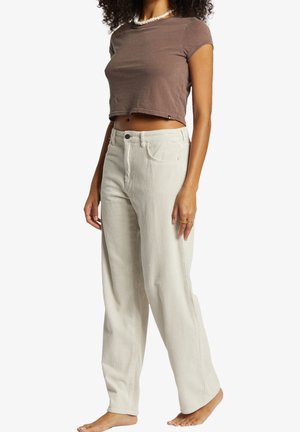 Frau trägt beige weite Cordhosen und ein kurzes braunes T-Shirt, steht barfuß vor weißem Hintergrund.