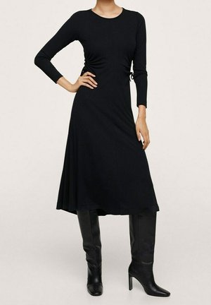 Mujer con un vestido midi negro de manga larga con lazos laterales y botas negras altas de tacón, de pie con una mano en la cadera.