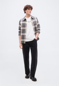 Νέος άνδρας με μπούκλες, φορώντας μία μπεζ και μαύρη καρό πουκάμισο πάνω από ένα λευκό μπλουζάκι με σχέδιο, μαύρο παντελόνι και μαύρα παπούτσια, στέκεται με το ένα χέρι στην τσέπη.