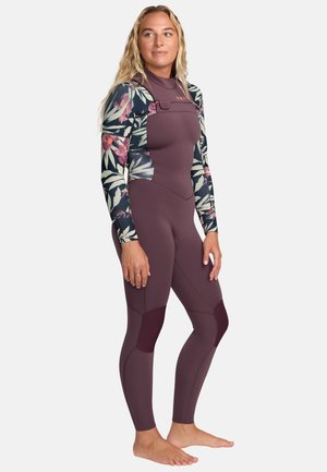 Kobieta stojąca boso, ubrana w kombinezon do nurkowania na całe ciało z kwiatowym wzorem na rękawach oraz jednolitym bordowym tułowiem i nogawkami.