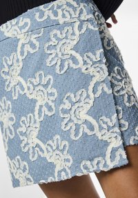 Denim mini-jumpe i lys blå, med hvite broderte blomstermønstre og en teksturert overflate med små prikker. Kanten er litt frynset.