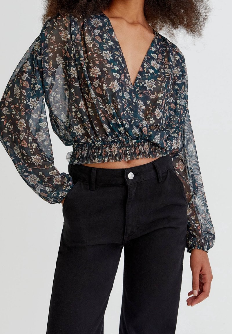 Femme portant un crop top transparent à manches longues à fleurs avec une taille élastique et un pantalon noir taille haute sur un fond uni.