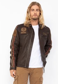 Giacca di pelle marrone con due tasche anteriori con zip, strisce laterali in ruggine e crema, e numerosi ricami sul petto e sulle maniche.