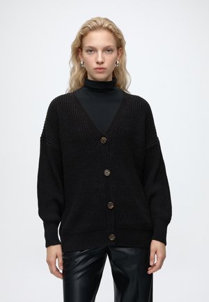 Blonde Frau mit silbernen Creolen, schwarzer, geknöpfter Strickjacke, schwarzem Rollkragenpullover und schwarzer Lederhose, stehend vor weißem Hintergrund.