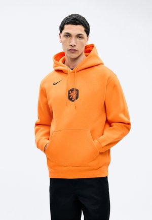 Homme portant un sweat à capuche orange vif avec un écusson de lion noir et le logo Nike, les mains dans la poche avant, debout devant un fond clair uni.