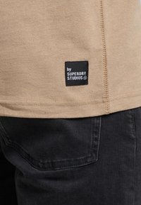 Camisa de algodón marrón clara con etiqueta negra que dice "by SUPERDRY STUDIOS." Presenta un dobladillo cosido. Jeans oscuros parcialmente visibles en el fondo.