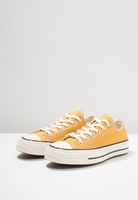 Gula canvas-sneakers med vita snören, rund tåhuvud och svart gummisula. Har förstärkta öglor och kontrasterande sömdetaljer.