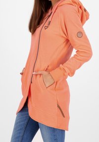 Hoodie rose avec fermeture éclair, poches latérales et détail en forme de cœur. Présente des poignets côtelés, des manches longues et une coupe décontractée.
