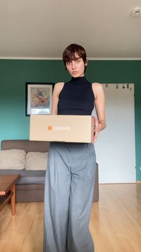 Caixa de cartão da Zalando segurada por uma pessoa vestindo uma blusa preta sem mangas e calças largas cinzas, com uma parede e um sofá verdes ao fundo.