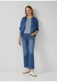 Kobieta stoi przy szarej ścianie, ubrana w niebieską bluzę na zamek, w paskowaną koszulę, niebieskie dżinsy i białe trampki, z jedną ręką w kieszeni.