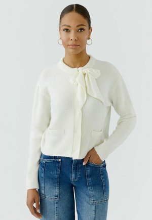 Vrouw met donker haar in een witte trui met strikdetail en blauwe spijkerbroek staat tegen een eenvoudige lichte achtergrond met één hand in de zak.