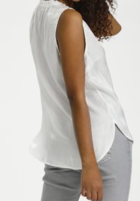 Biała bezrękawnikowa bluzka o miękkiej, lekkiej teksturze, z marszczonym dekoltem i zaokrąglonym dolnym brzegiem. Noszona z szarymi spodniami.