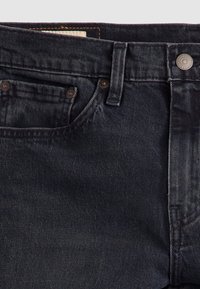 Gros plan d'un jean en denim noir montrant la poche avant, la boucle de ceinture et la fermeture par bouton en métal.