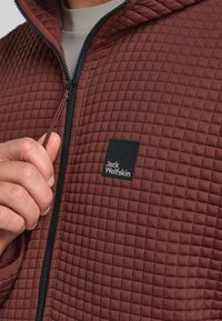 Burgunderfarbene gesteppte Jacke mit Reißverschluss, einem schwarzen Logopatch und einem strukturierten Stoff. Mit Kapuze und verstellbarem Kordelzug.