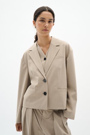 Žena s tmavými vlasy staženými dozadu, oblečená v béžovém nadměrném blejzru a odpovídajících plisovaných kalhotách, na bílém jednobarevném pozadí.