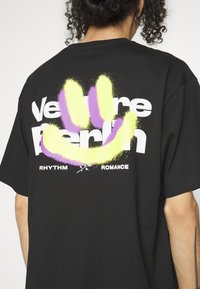 Zwarte katoenen oversized t-shirt met witte tekst "Venture Berlin" en "Rhythm Romance," voorzien van een kleurrijk gesprayde smiley-ontwerp.