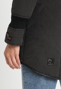 Giubbotto imbottito nero con polsini a coste; presenta un'etichetta in tessuto con il logo "KHUJO" e una leggera lucentezza; abbinato a jeans blu.