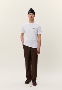 Jauns vīrietis, tērpies baltā kreklā ar apdruku, tumši brūnās biksēs, melnā cepurē un tumšās kurpēs, stāvot ar vienu roku kabatā.