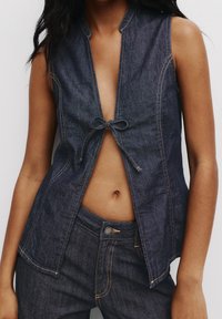 Gilet en denim avec un décolleté profond, noué à la taille avec un nœud. Présente des coutures apparentes et une légère forme évasée. Couleur bleu foncé.