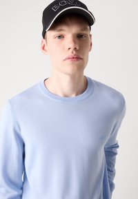 Suéter azul-claro de gola redonda com uma textura suave e punhos canelados, combinado com um boné preto com a palavra "BOSS" em bordado branco.