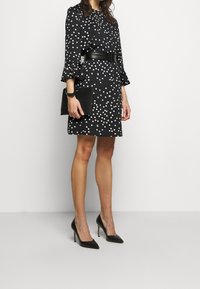 Mustapuku, jossa on valkoisia pilkkuja, volangihihat, kireä musta vyö, yhdistettynä mustiin kärkitossuihin ja mustaan käsilaukkuun.