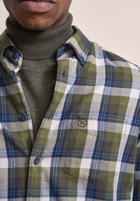 Grün, blau und weiße karierte Flanellbluse mit einem Button-Down-Kragen, ausgestattet mit einer Brusttasche und einem gerippten olivgrünen Rollkragenpullover darunter.