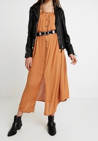 Kvinna i en knappad orange maxikjol med ett svart bälte, svart skinnjacka och svarta ankelskor som står mot en vit bakgrund.