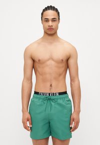Pantaloni de baie verzi cu un talie elastică, cu un logo negru. Textură netedă, design până la genunchi și șnur în față pentru ajustare.