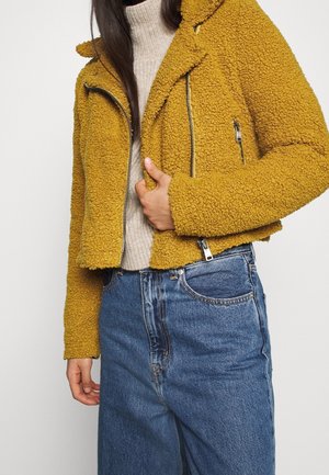 Personne portant une veste zippée jaune moutarde texturée sur un pull à col roulé beige, associée à un jean bleu taille haute.