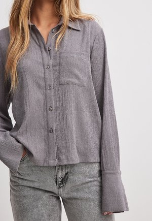 Frau trägt ein strukturiertes graues Hemd mit Knöpfen, einer Brusttasche und langen Ärmeln, kombiniert mit hellgrauen, gewaschenen Jeans.