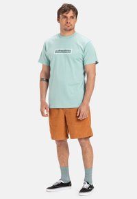 Světle modré bavlněné tričko s kostkovaným logem, spojené s oranžovými manšestrovými šortkami. Černé tenisky a tyrkysové ponožky doplňují outfit.