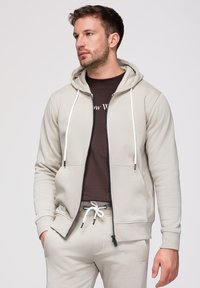 Homme portant un sweat à capuche beige clair zippé et un pantalon de jogging assorti, debout les mains dans les poches, regardant vers la gauche, avec un t-shirt marron foncé en dessous.