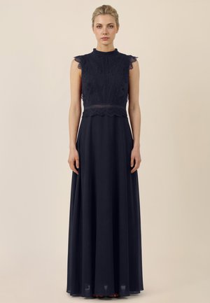 Frau trägt ein ärmelloses schwarzes Kleid aus Spitze mit hohem Kragen und bodenlangem, fließendem Rock, stehend vor einem einfarbigen beigen Hintergrund.