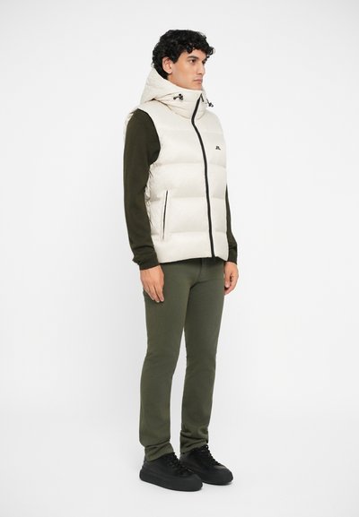 Puffer vest in light beige with hood, black zippers, and two side pockets. Worn over a dark green long-sleeve top and olive green pants.