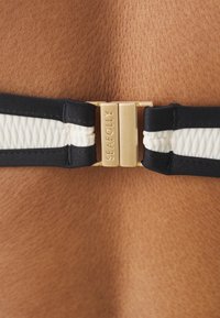 Zwart-witte elastische band met een gouden metalen sluiting waarop de gegraveerde tekst "SEAFOLLY" staat. De textuur lijkt glad aan tegen de huid.
