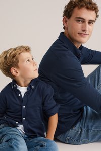 Tumši zili garie piedurkņu krekli ar pogājamām apkaklēm, ko valkā vīrietis un zēns. Zēna krekla piedurknes ir uzrullētas. Abi valkā džinsus.