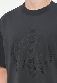 Tumši pelēks kokvilnas T-krekls ar izcelta grafiskā attēla printu, uz kura redzams regbija bumbas un lauru lapu attēls, kā arī teksts "All Blacks" un "Originals."
