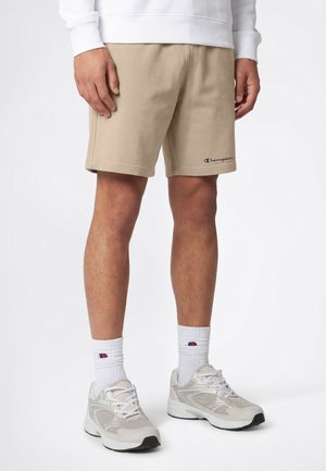 Mężczyzna w beżowych spodenkach Champion, białych skarpetkach z logo Champion oraz białych sportowych butach, stojący na tle jednolitego białego tła.