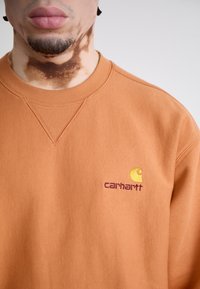 Orange sweatshirt med ribbet halsudskæring, som har et broderet logo i gul og bordeaux. Fremstillet af bomuldsstof med en blød tekstur.