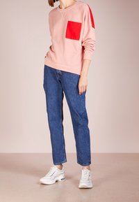 Sudadera rosa con un bolsillo parche rojo, combinada con vaqueros azules y zapatillas blancas. Cuenta con un corte holgado y un diseño de cuello redondo.